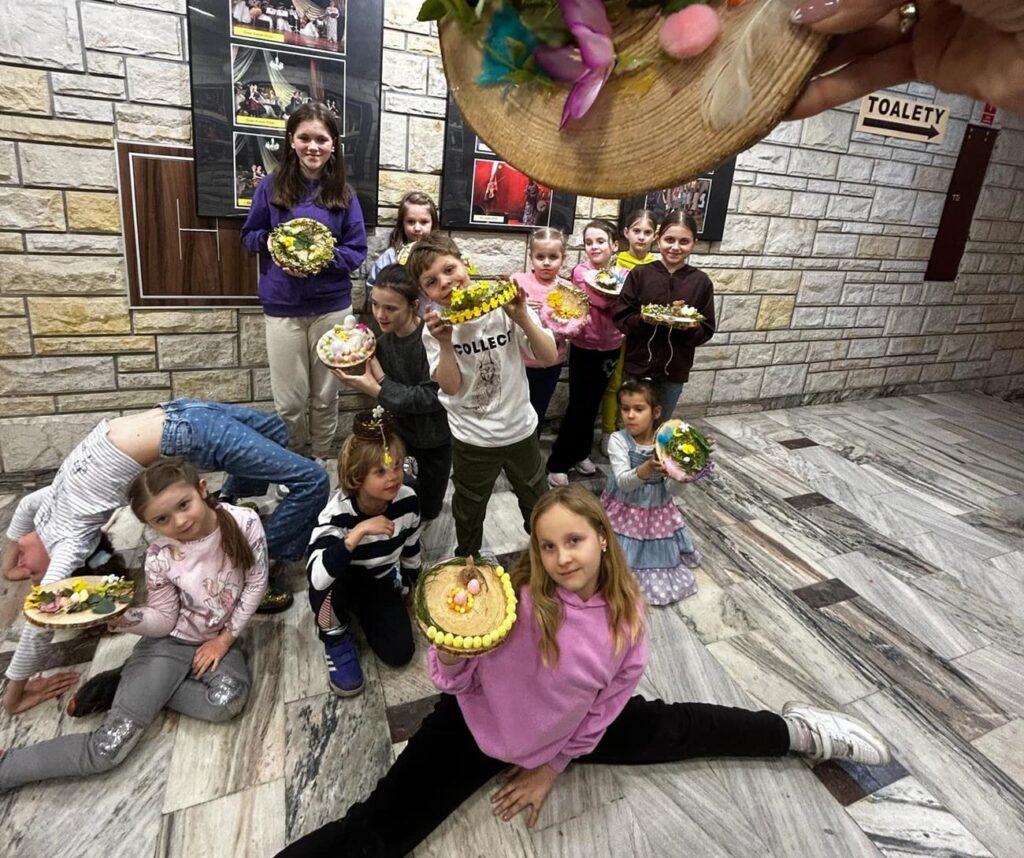 Grupa kilkunastu dzieci w kolorowych ubraniach uśmiechają się i prezentują swoje prace/stroiki wielkanocne. Niektórzy stoją, inni kucają lub wyginają się w różnych pozycjach. Pomieszczenie oświetlone ciepłym światłem.