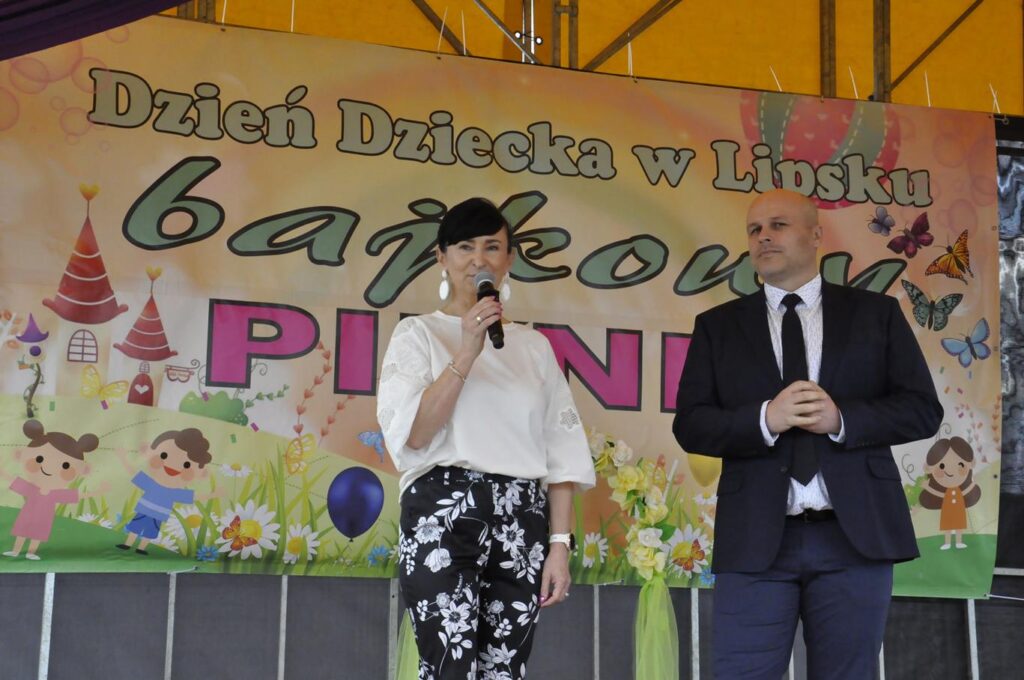 Kobieta w białej koszulce trzyma mikrofon. Obok niej stoi mężczyzna w czarnej marynarce. Za nimi kolorowy baner promujący wydarzenie.