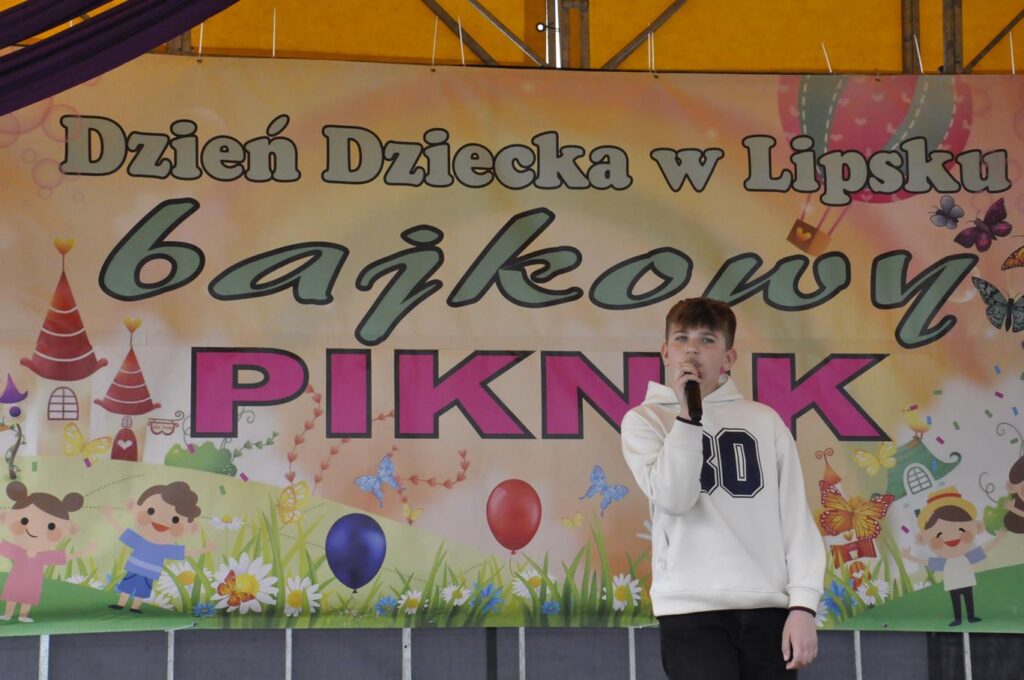 Chłopiec w białej bluzie z czarnym numerem śpiewa do mikrofonu. Za nim wisi baner z nazwą wydarzenia.