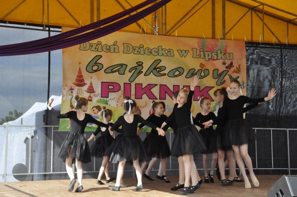 Dziewczynki w czarnych sukienkach tańczą na scenie. Za nimi baner z nazwą wydarzenia.