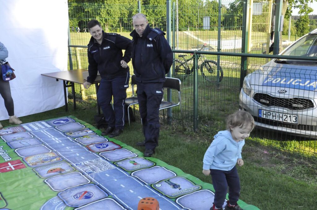 Małe dziecko skacze po macie edukacyjnej. Przygląda się mu dwoje funkcjonariuszy policji. Niedaleko stoi radiowóz. Na dalszym planie ściana lasu.