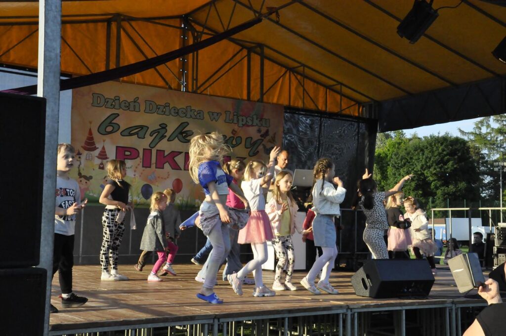 Grupa dzieci w kolorowych ubraniach skacze po scenie plenerowej. Za nimi DJ przy konsoli. Pogodny dzień.