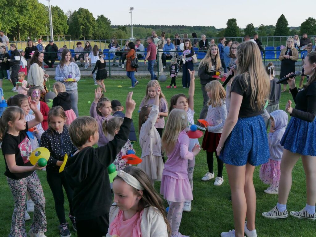 Boisko. Grupa dzieci podnosi palce do grup. obok stoją dwie dziewczyny w granatowych sukienkach. Trzymają mikrofony. W dalszym planie grupa ludzi siedzi na stadionowych niebieskich krzesełkach.