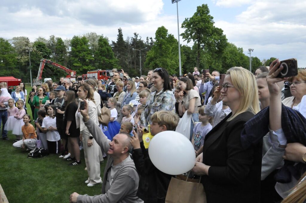 Tłum publiczności w zwykłych ubraniach. W tle wóz strażacki i ściana lasu.
