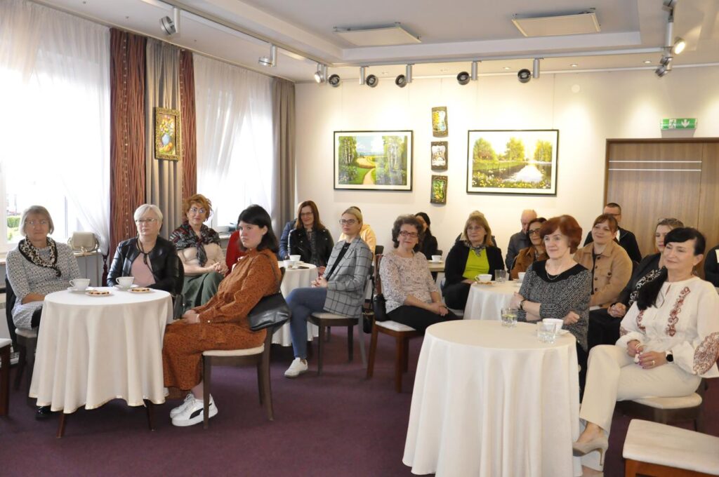Duża grupa ludzi w zwyczajnych strojach siedzących przy okrągłych stolikach przykrytych białymi obrusami. Na stolikach stoją filiżanki. W tle obrazy krajobrazów na jasnej ścianie. Pomieszczenie ciepło oświetlone