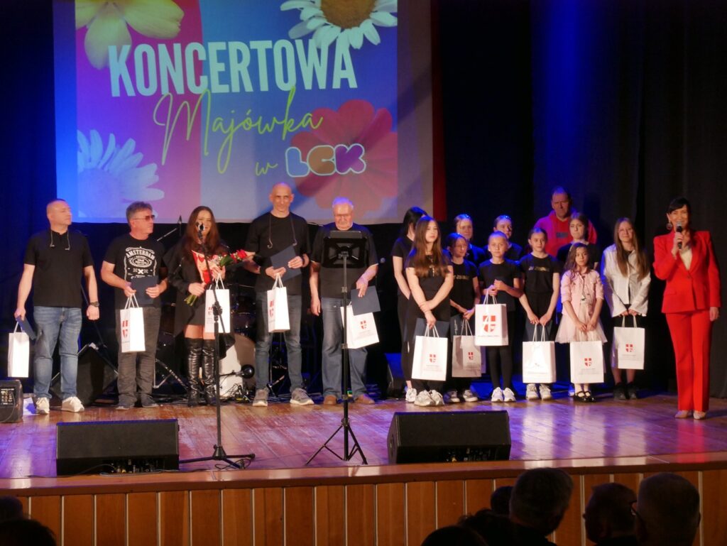 Dorośli i dzieci stoją na scenie. Wszyscy trzymają białe torby z herbem miejski. Kobieta w czerwonym stroju trzyma mikrofon. Nad nimi wyświetlony baner promujący wydarzenie.