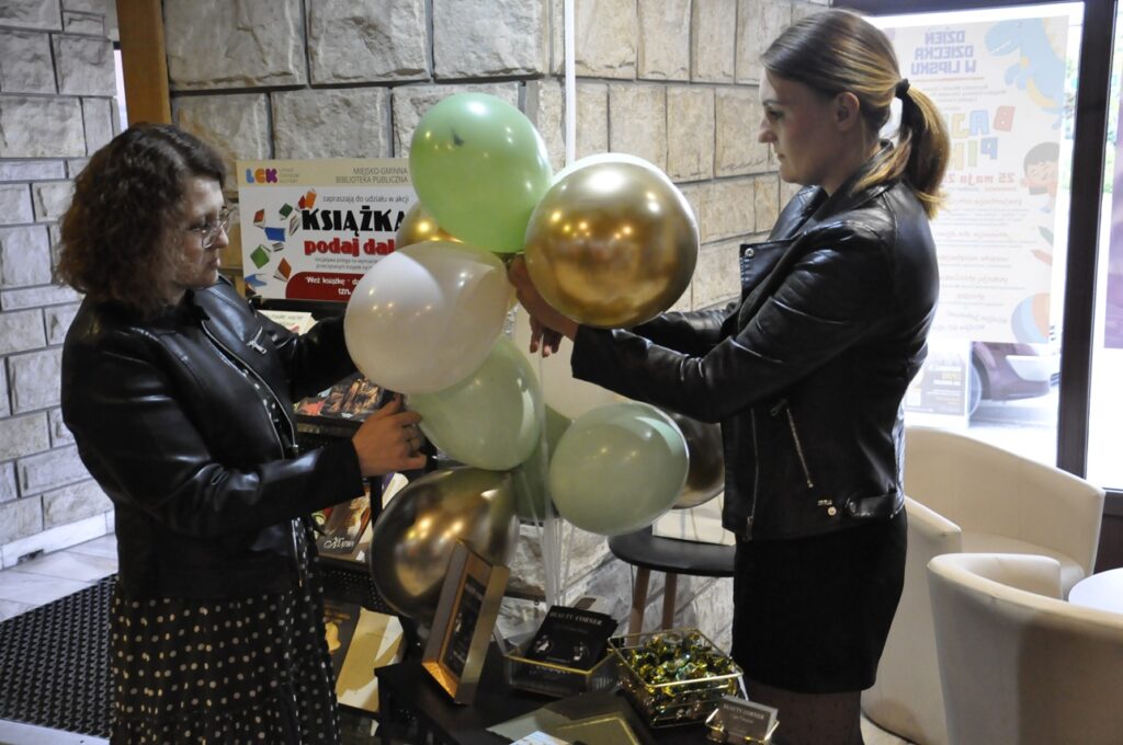 Dwie kobiety w czarnych kurtkach trzymają złote, białe i jasnozielone balony. Pomieszczenie jasno oświetlone.