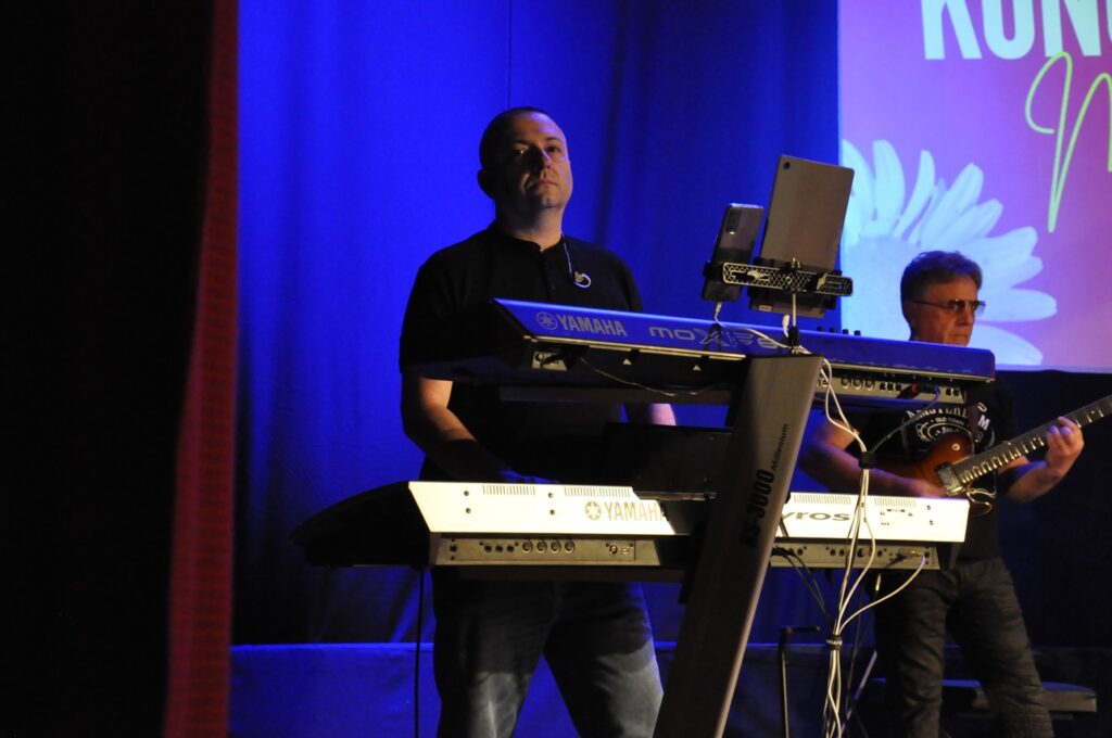 Mężczyzna w czarnej koszulce stoi przy keyboardzie. Tło niebieskie z fragmentem wyświetlanego banera wydarzenia.