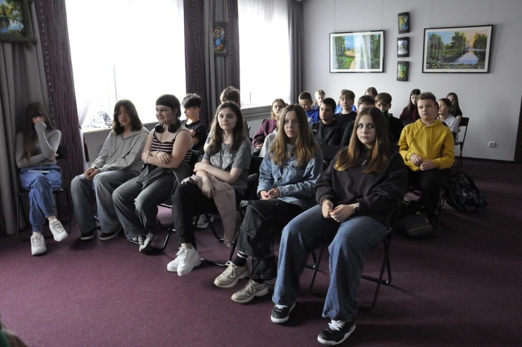 Młodzież w zwykłych ubraniach siedzi na krzesłach. Za nimi biała ściana, na której wiszą obrazy natury. Pomieszczenie oświetlone dziennym światłem.