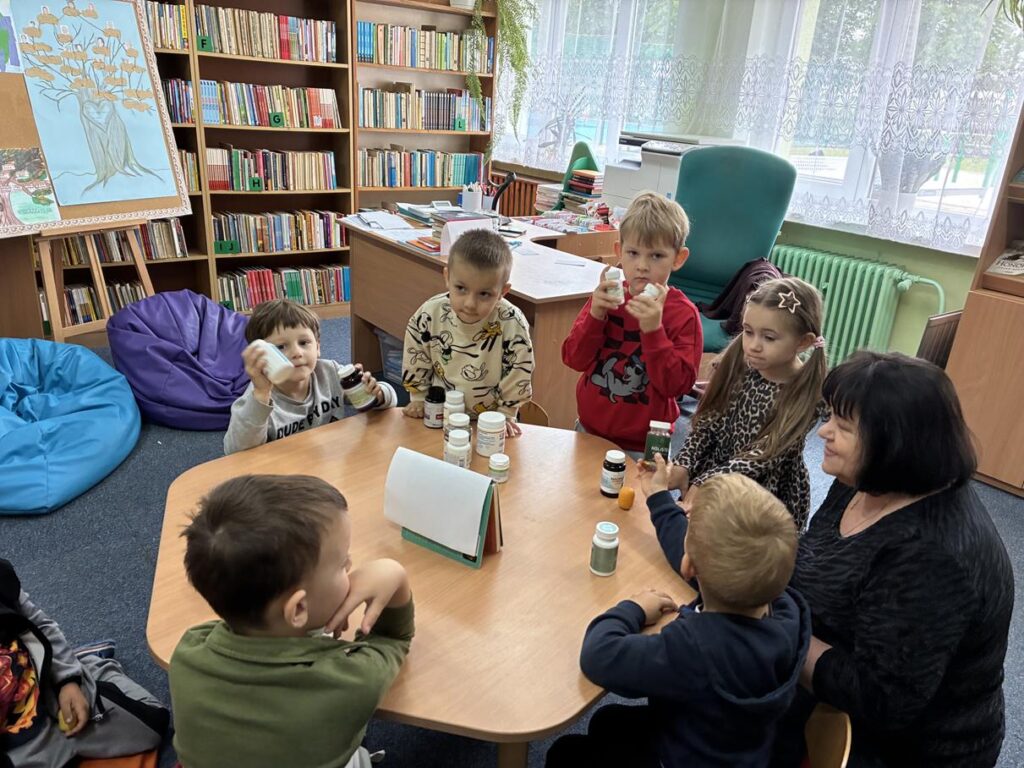 Kilkoro dzieci i kobieta siedzą przy stoliku na którym stoją plastikowe pojemniki. Wokół nich biurko i półki z książkami.