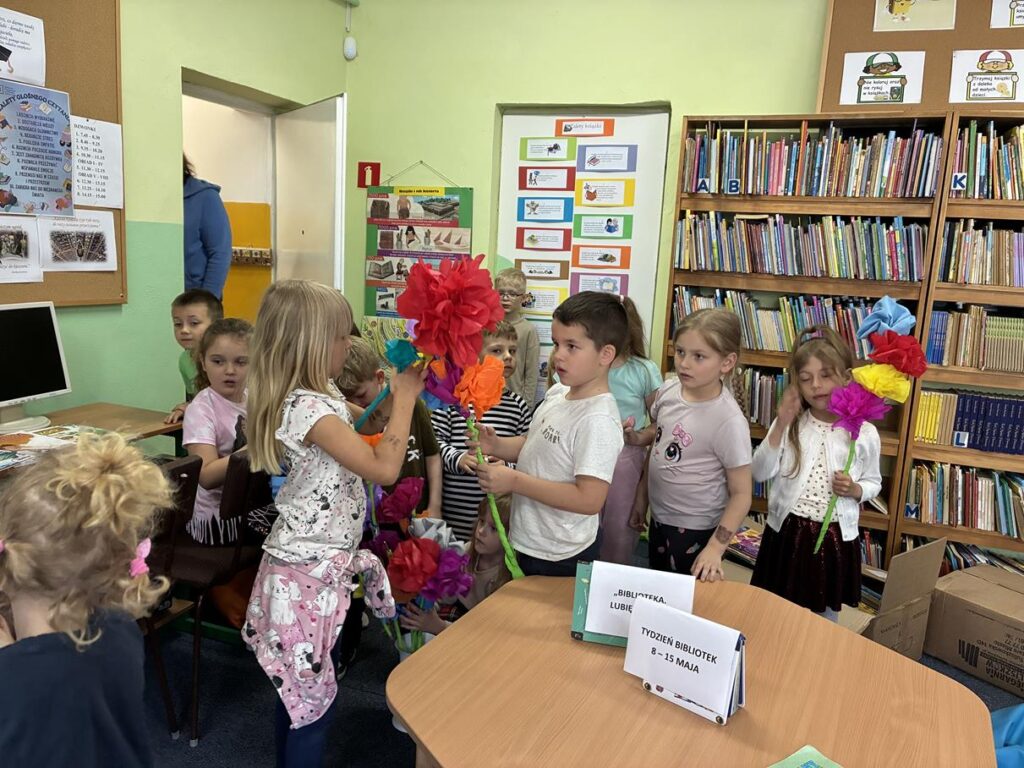 Kilkoro dzieci trzyma kolorowe prace, wiosenne kwiaty. Pomieszczenie biblioteczne.