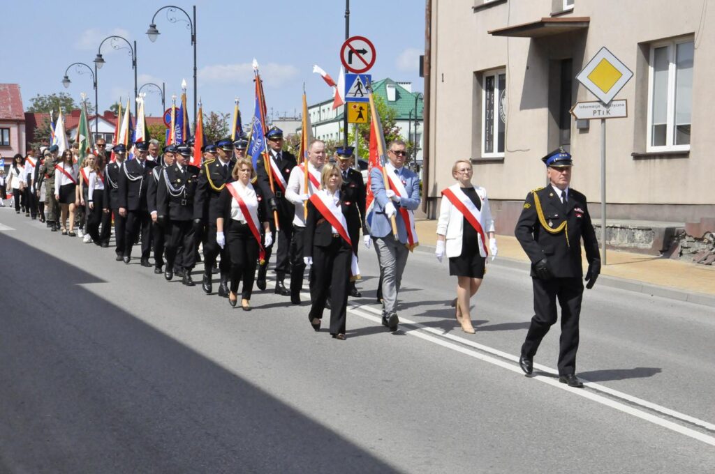 Ulica miasta. Kilkadziesiąt osób w oficjalnych, uroczystych i służbowych strojach mazeruje. Niektórzy trzymają sztandary. Pogodny dzień.