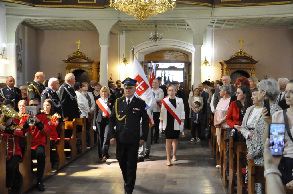 Wnętrze kościoła. Pogodne oświetlenie. Grupa osób w oficjalnych strojach na czele z mężczyzną w mundurze strażackim idą przez środek kościoła. Przyglądają się temu osoby stojące między ławkami.