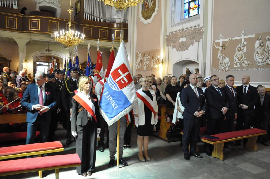 Wnętrze kościoła. Ciepłe oświetlenie. Uczestnicy wydarzenia stoją między ławkami. Niektórzy trzymają sztandary.