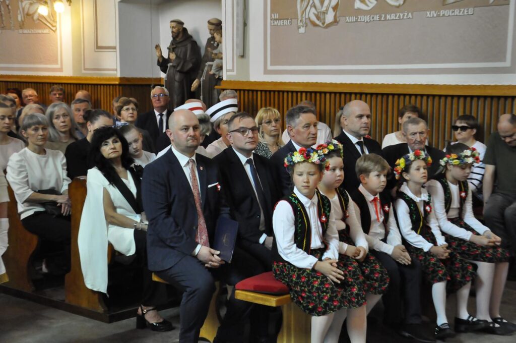 Ludzie w oficjalnych i służbowych strojach siędzą w ławkach. Kilka dzieci w pierwszej ławce w strojach ludowych. Ciepłe oświetlenie.