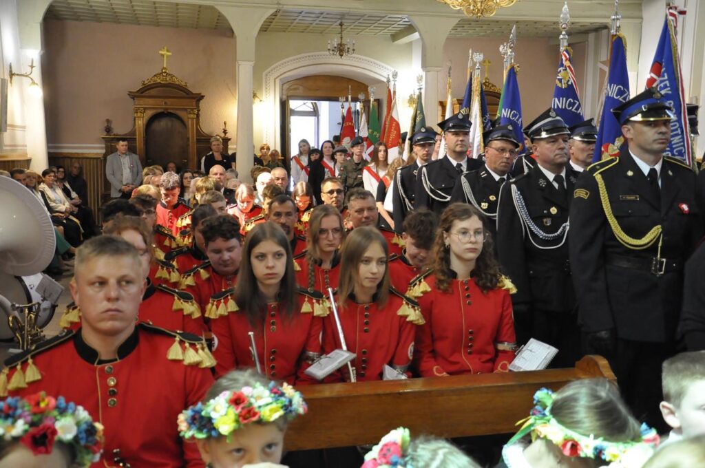 Grupa osób w czerwonych strojach i z instrumentami muzycznymi siedzi w ławkach. Pozostali w strażackich mundurach stoją przy sztandarach.