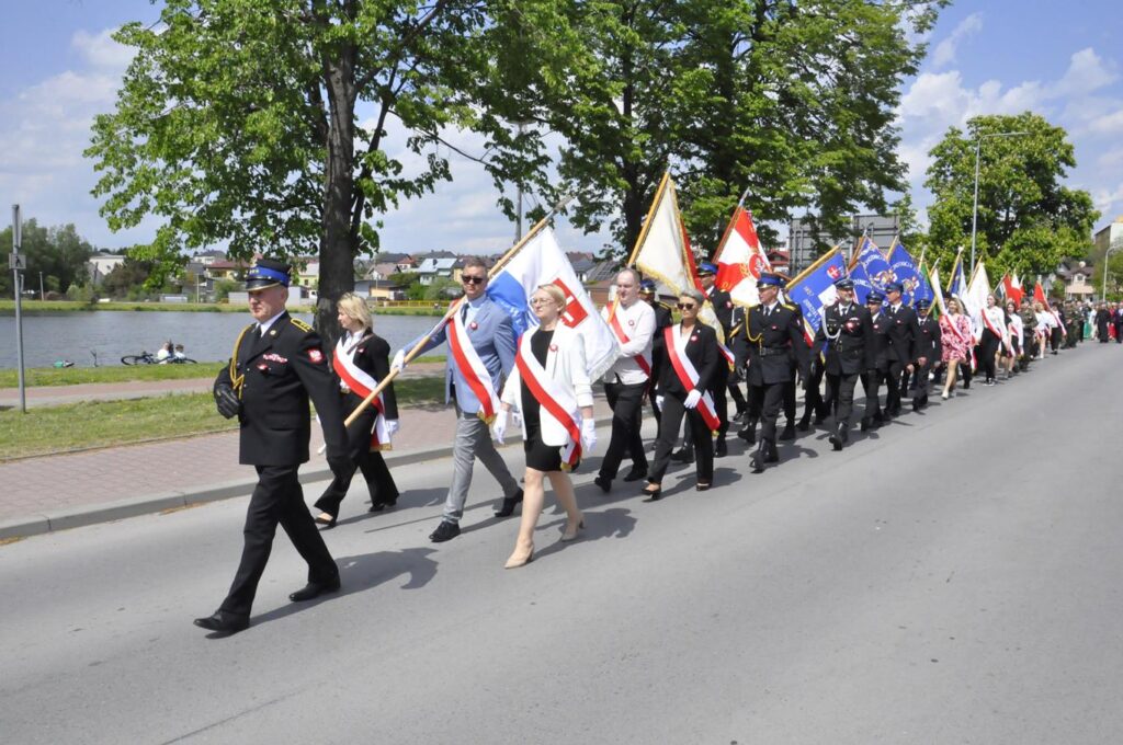 Grupa odświętnie ubranych osób maszeruje ze sztandarami przez miasto. Pogodny dzień. W tle duży zbiornik wodny.