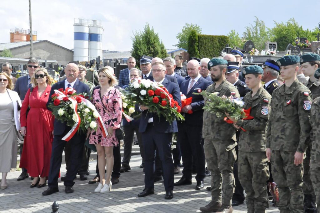 Grupa odświętnie ubranych osób, niektórzy w mundurach służbowych stoją. Kilka osób trzyma biało-czerwone wiązanki. W tle nagrobki.