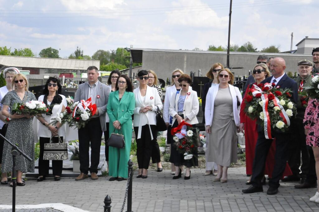 Kilkanaście osób w odświętnych strojach stoi w otoczeni nagrobków. Niektórzy trzymają biało-czerwone wiązanki. Pogodny dzień.