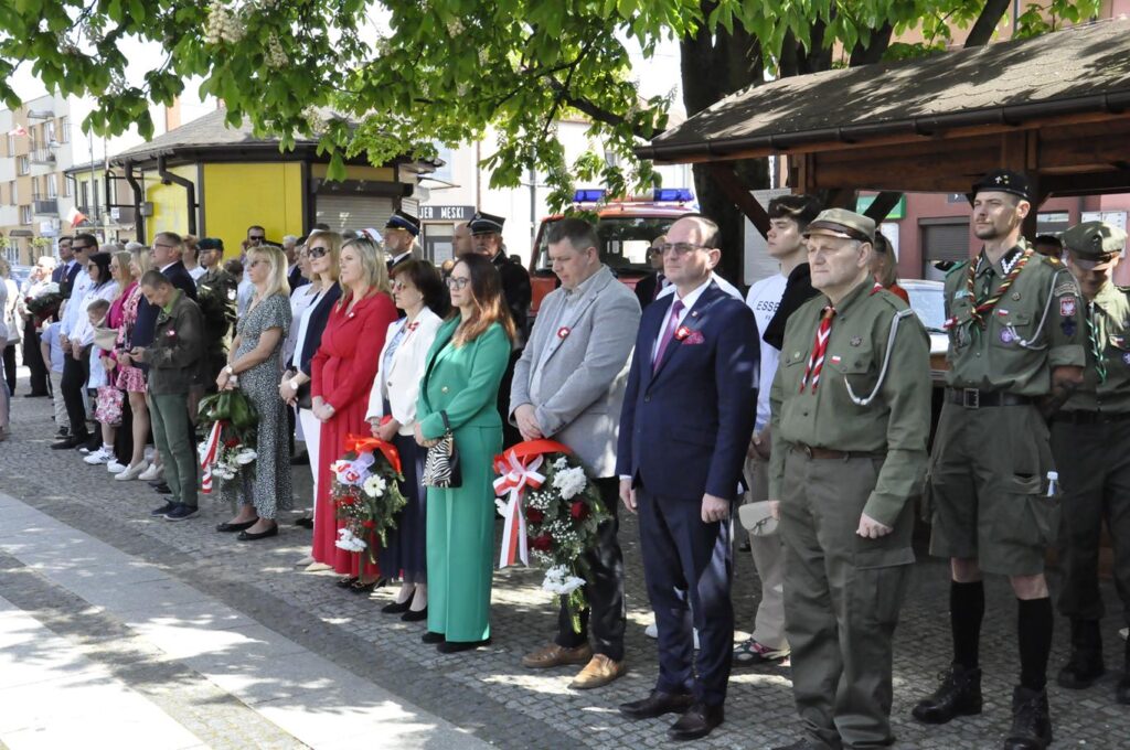 Kilkadziesiąt osób w kolorowych oficjalnych strojach, w tym w mundurach stoi w cieniu drzewa . Niektórzy trzymają biało-czerwone wiązanki kwiatów. Słoneczna pogoda.