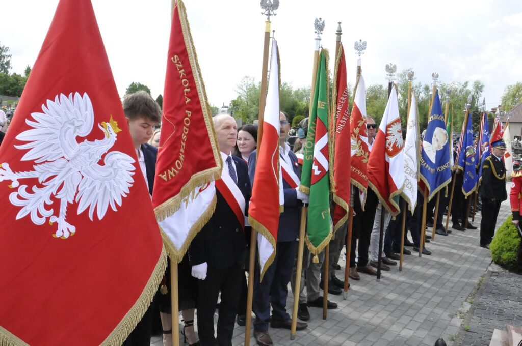 Kilkanaście osób stoi w rzędzie. Niektóry trzymają kolorowe sztandary. Pogodny dzień.