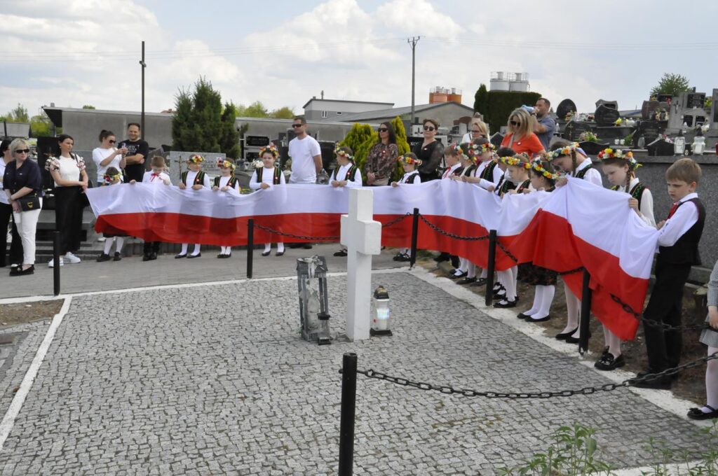 Dzieci w ludowych strojach trzymają dużą szarfę w barwach narodowych. Wokół nagrobki. Pogodny dzień.