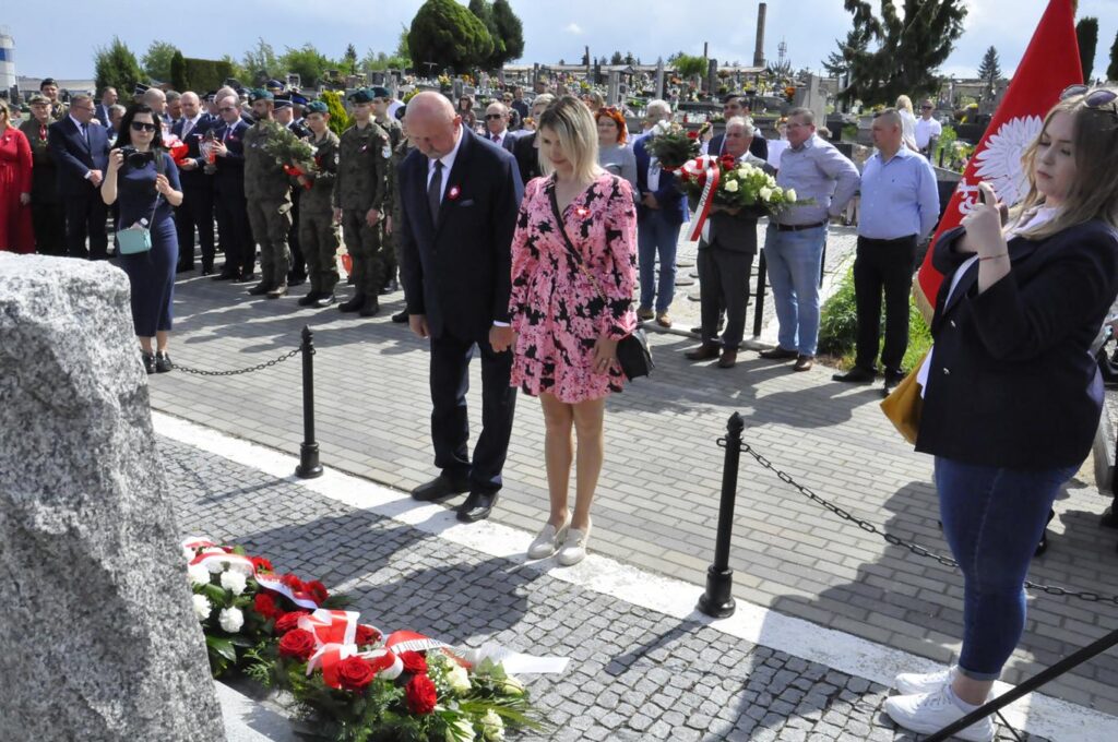 Mężczyzna w ciemnym, granatowych garniturze i kobieta w różowej sukience pochylają głowę przy złożonych wiązankach kwiatów. Za nim kilkanaście osób, niektórzy trzymają wiązanki kwiatów.. Pogodny dzień.