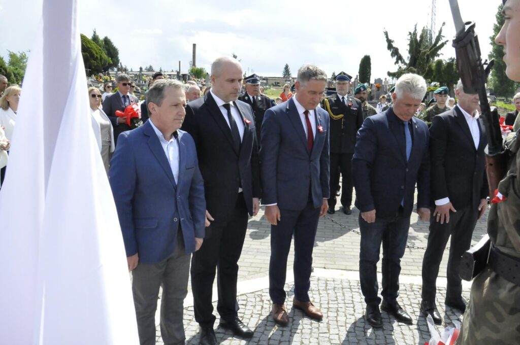 Pięciu mężczyzn w garniturach pochyla głowę, za nimi więcej osób w oficjalnych strojach. Wokół nagrobki. Pogodny dzień.