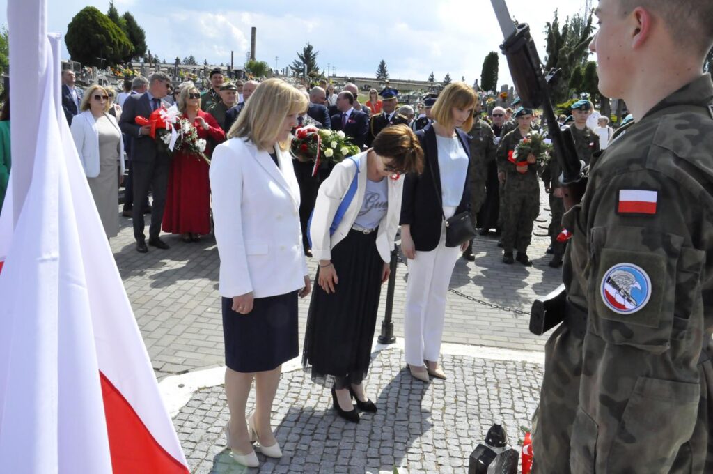 Trzy kobiety w biało-czarnych strojach pochyla się przy złożonymi zniczami. Za nimi kilkanaście osób w oficjalnych strojach. Niektórzy trzymają wiązanki biało-czerwonych kwiatów. Pogodny dzień.