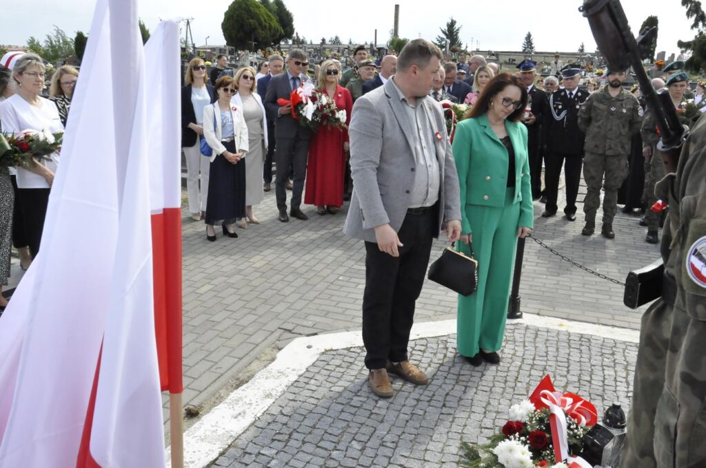 Mężczyzna w szarej marynarce i kobieta w zielonym żakiecie pochylają głowy przy złożonej wiązance biało-czerwonych kwiatów. Mężczyzna w ciemnym, granatowych garniturze pochyla głowę przy złożonej wiązance kwiatów. Za nim kilkanaście osób, niektórzy trzymają sztandary. Pogodny dzień.