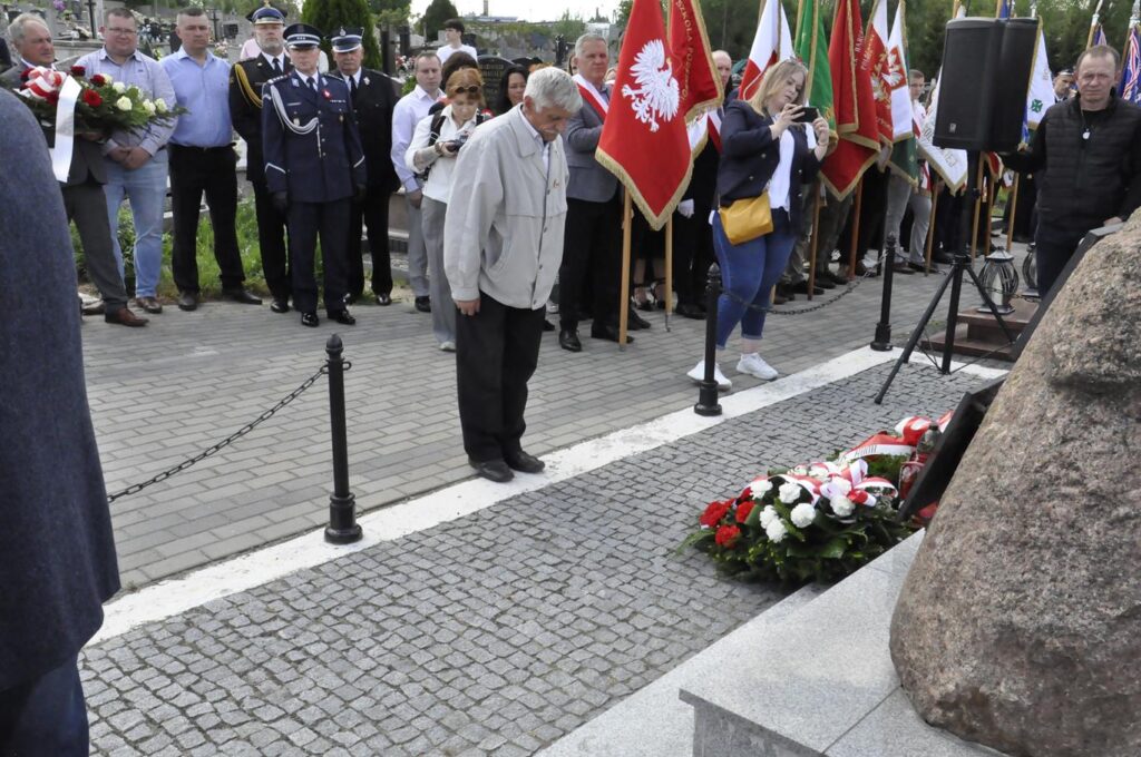 Mężczyzna w szarej kurtce pochyla głowę przy złożonej wiązance kwiatów. Za nim kilkanaście osób, niektórzy trzymają sztandary i wiązanki. Pogodny dzień.