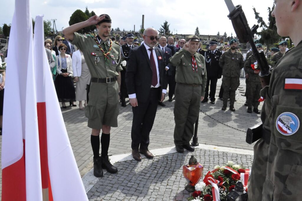 Trzech mężczyzn, jeden w garniturze, pozostali w mundurach harcerskich. Składają hołd nad złożonymi wiązankami. Za nim kilkanaście osób w oficjalnych i służbowych strojach. Pogodny dzień.