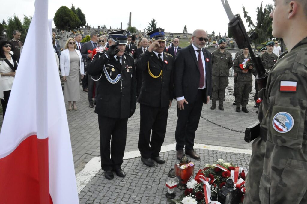 Trzech mężczyzn, jeden w czarnej marynarce, pozostali w mundurach strażackich składają hołd przy złożonych wiązankach i zniczach. Za nim kilkanaście osób w odświętnych strojach. Pogodny dzień.