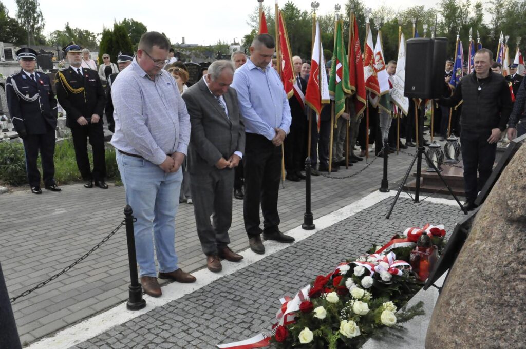 Trzech mężczyzn w jasnych koszulach pochyla głowę nad złożonymi wiązankami kwiatów. Za nim kilkanaście osób, niektórzy trzymają sztandary. Pogodny dzień.