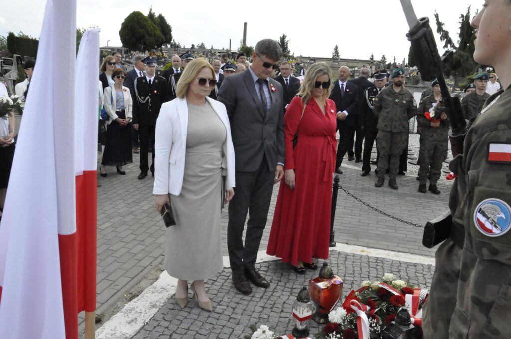 Mężczyzna w szarym garniturze i dwie kobiety, jedna w czerwonej sukience, druga w białej marynarce pochylają głowy nad złożonymi wiązankami. Za nim kilkanaście osób w odświętnych strojach. Pogodny dzień.