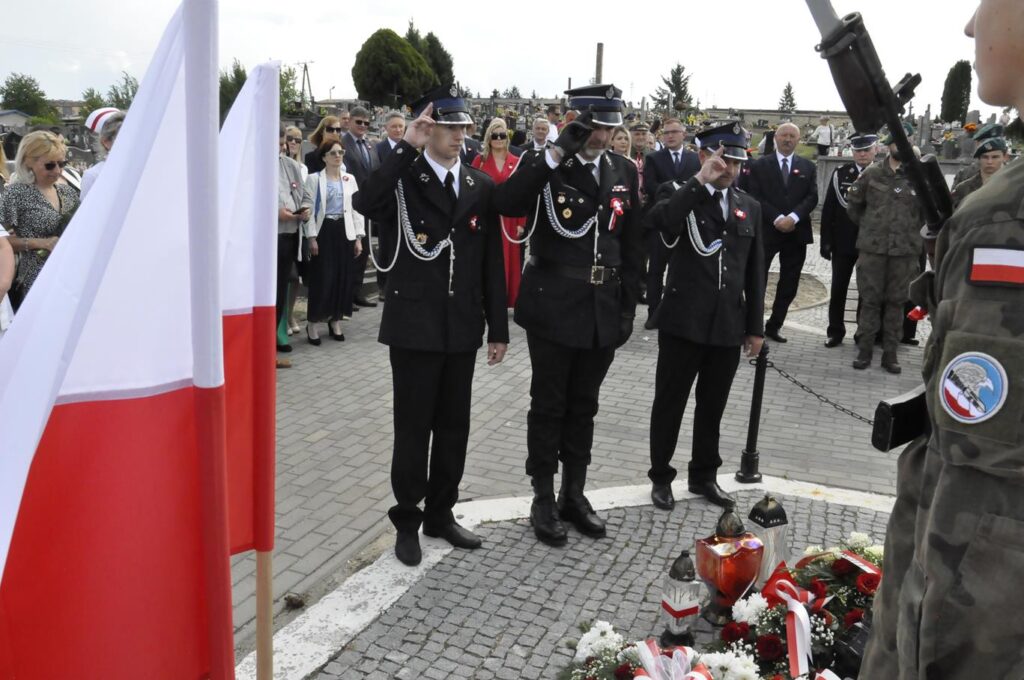 Trzech mężczyzn w strażackich mundurach składa hołd nad złożonymi wiązankami biało-czerwonych kwiatów. Za nim kilkanaście osób w odświętnych strojach. Pogodny dzień.