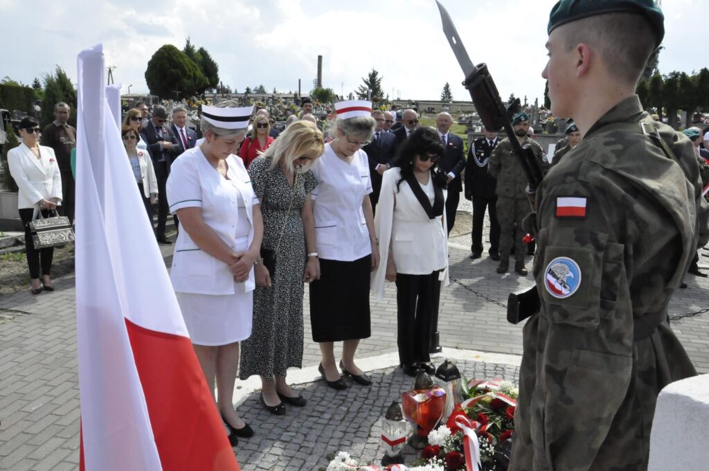 Cztery kobiety pochylają głowę. Dwie mają czepki pielęgniarskie. Za nim kilkanaście osób w odświętnych strojachy. Pogodny dzień.