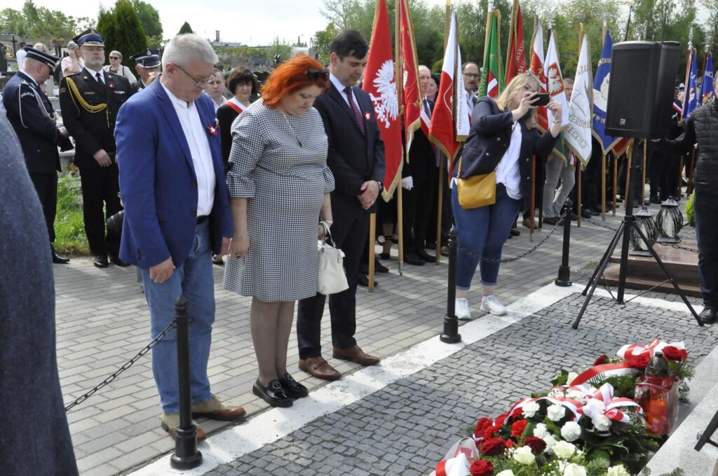 Dwóch mężczyzn w marynarkach i kobieta w sukience drobną biało-czarną kratkę pochylają głowę nad złożonymi wiązankami kwiatów. Za nim kilkanaście osób, niektórzy trzymają sztandary. Pogodny dzień.