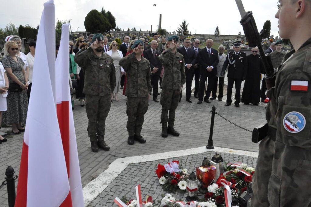 Trzech młodych mężczyzn w strzeleckich mundurach składa hołd przed złożonymi wiązankami kwiatów. Za nim kilkanaście osób w oficjalnych strojach. Pogodny dzień.