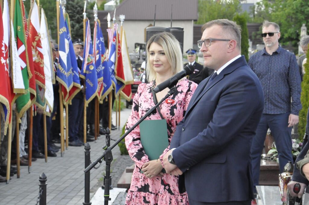 Mężczyzna w garniturze przemawia do mikrofonu na statywie. Obok niego stoi kobieta w różowej sukience. W tle ludzie ze sztandarami. Pogodny dzień.