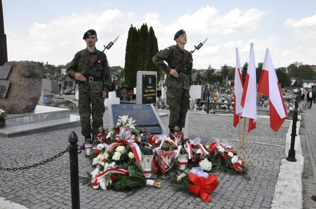 Dwóch mężczyzn w mundurach strzeleckich trzyma wartę nad pomnikiem, przy którym leżą biało-czerwone wiązanki kwiatów i zapalone znicze. Obok statyw z trzema flagami w barwach narodowych. Dalej nagrobki. Pogodny dzień.