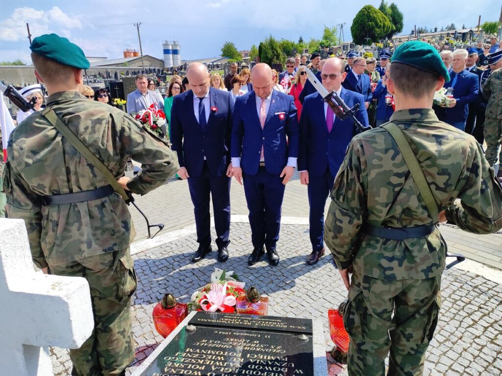 Trzech mężczyzna w śliwkowych garniturach pochyla głowę nad złożonymi wiązankami biało-czerwonych kwiatów. Za nim kilkanaście osób w kolorowych odświętnych strojach. Pogodny dzień.