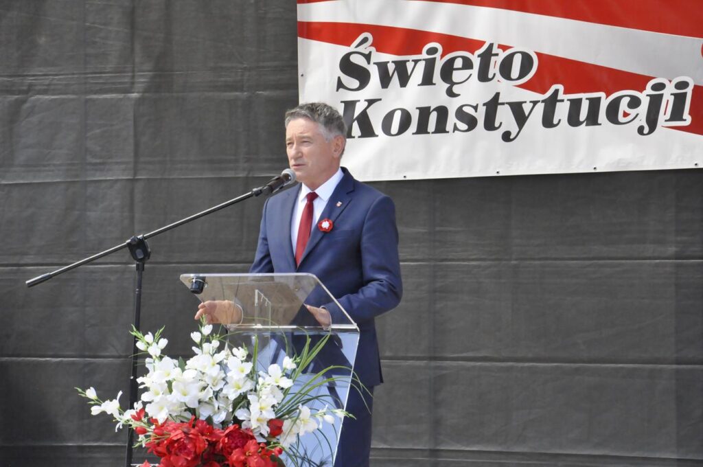 Mężczyzna w śliwkowym garniturze stoi za mównicą i mówi do mikrofonu na statywie. W tle biało czerwony baner z nazwą uroczystości.