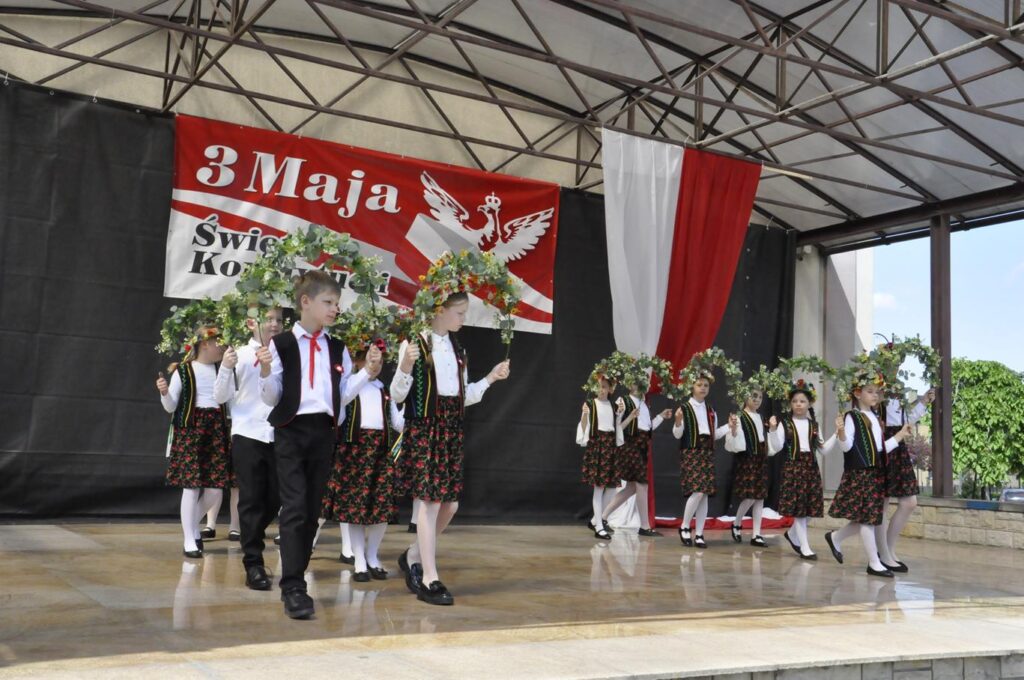 Scena plenerowa. Kilkanaścioro dzieci w strojach ludowych wznosi kolorowe, kwiatowe pałąki. W tle biało-czerwone dekoracje.