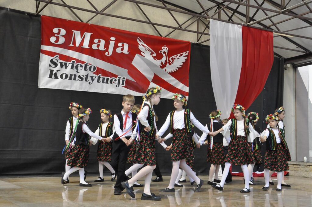 Scena plenerowa. Kilkanaścioro dzieci w strojach ludowych przechadza się po scenie. W tle biało-czerwone dekoracje.