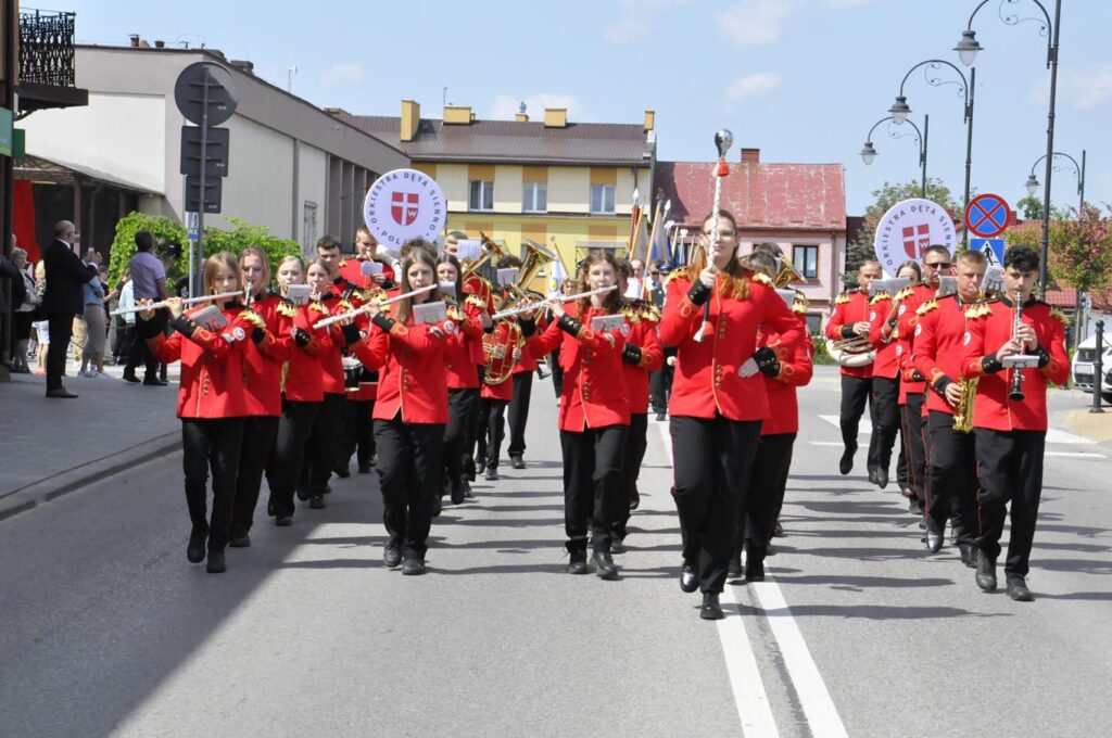 Ulica miasta. Kilkanaście osób w czerwonych, oficjalnych strojach idzie ulicą. Wszyscy trzymają instrumenty muzyczne. Pogodny dzień.