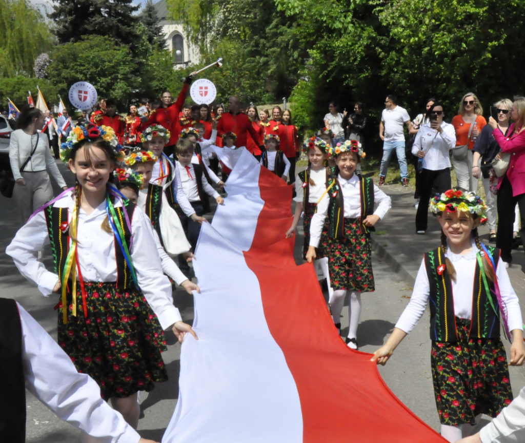Ulica miasta. Uśmiechnięte dzieci w ludowych strojach niosą flagę z barwami narodowymi. Pogodny dzień.