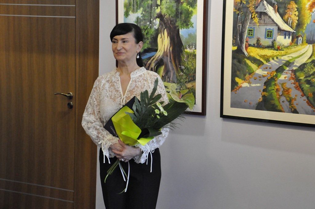 Kobieta w białej koronkowej koszuli i czarnymi włosami spiętymi w warkocz trzyma bukiet kwiatów. W tle obrazy natury na białej ścianie.