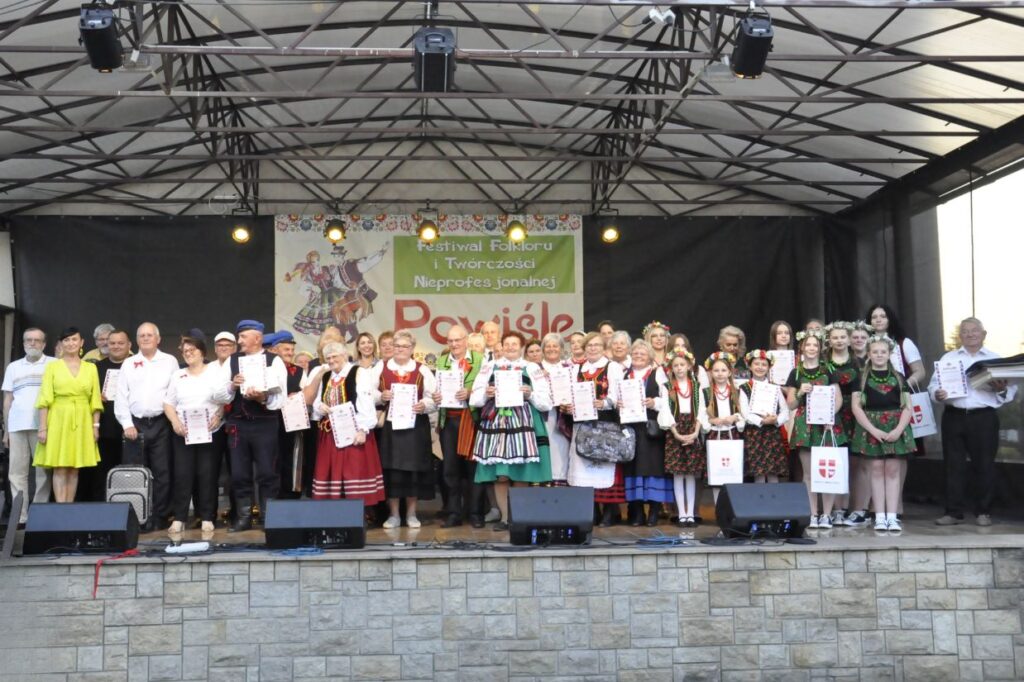 Liczna grupa dorosłych, młodzieży, dzieci i dorosłych pozuje do wspólnego zdjęcia na cenie plenerowej. Większość osób w strojach ludowych. Niektórzy trzymają dyplomy, inni statuetki. Tło czarno-granatowe. Nad nimi baner z nazwą wydarzenia.