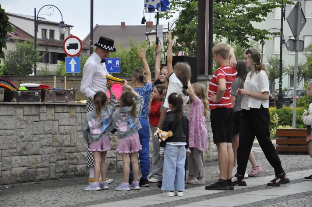 Grupa dzieci stoi obok cyrkowca przy scenie plenerowej. Niektóre dzieci podnoszą rękę do góry.